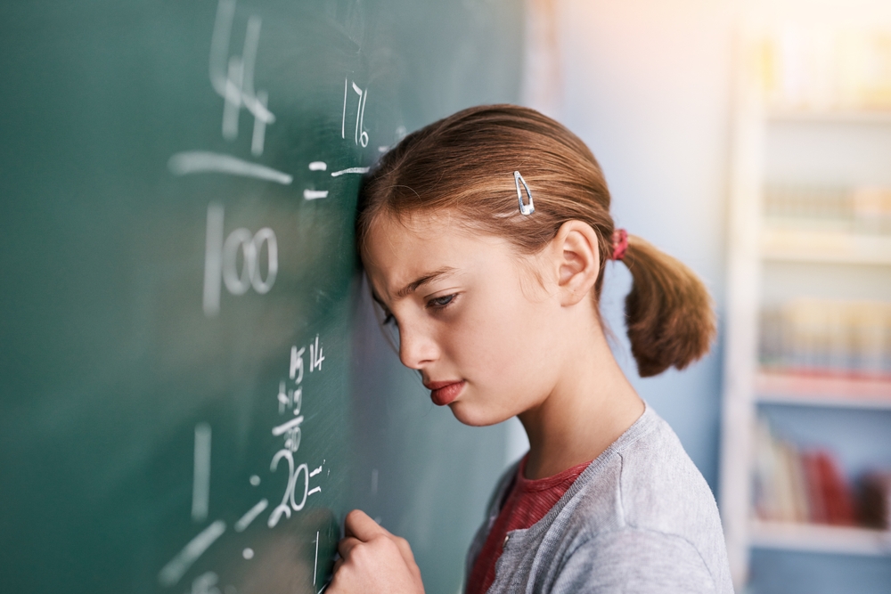 A young girl with a ponytail, resting her head against a blackboard while holding a piece of white chalk, looking perplexed.