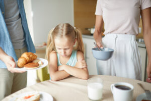 Blond-haired girl in green tank top with two ponytails sitting at the table with her arms crossed while mom and dad are offering two different plates of food.