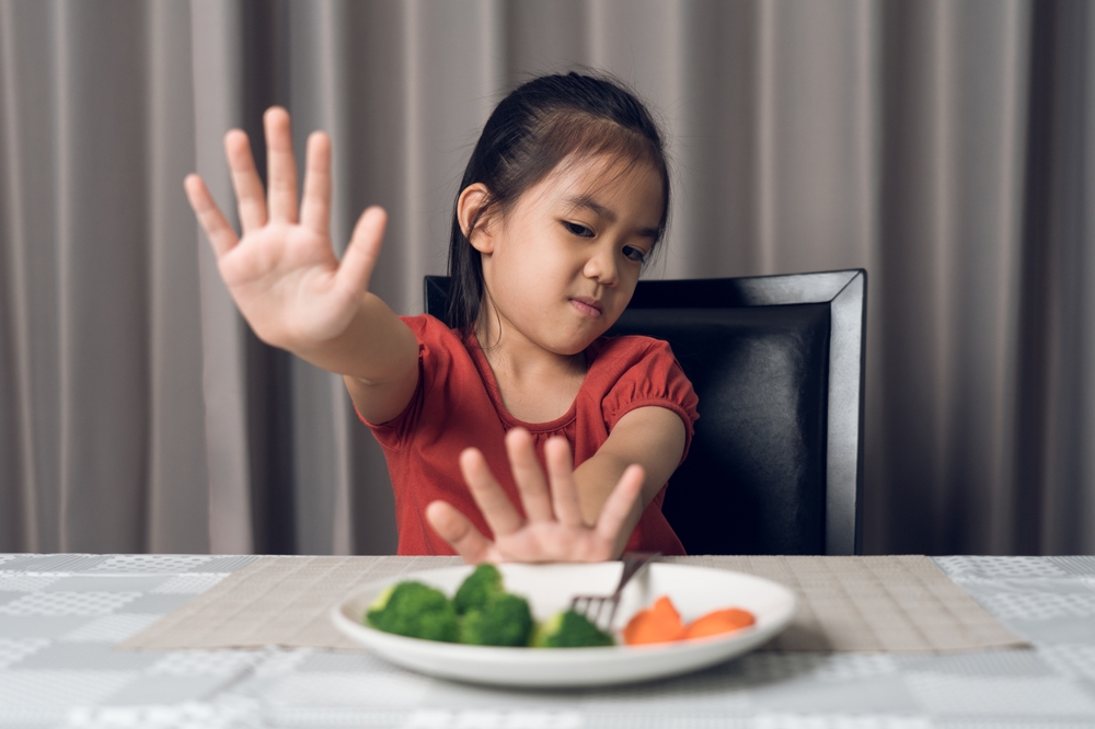 A young asian girl in a red shirt putting her hands up in refusal of the plate of vegetables sitting in front of her at the dinner table.