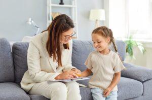 A therapist in a white suit, holding a clipboard and wearing glasses, working with a young girl sitting on a sofa wearing a cream t-shirt and jeans.