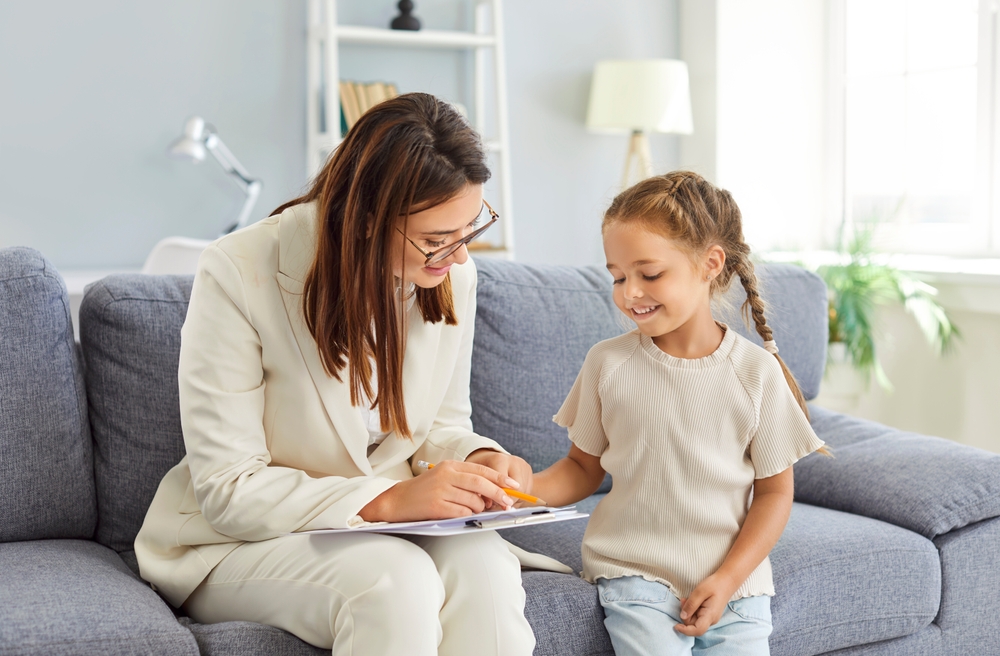 A therapist in a white suit, holding a clipboard and wearing glasses, working with a young girl sitting on a sofa wearing a cream t-shirt and jeans.