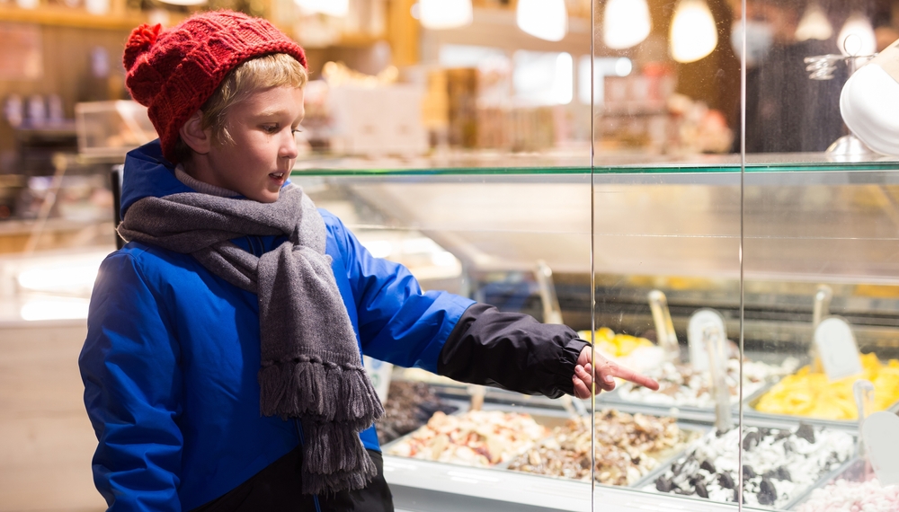 A blond-haired boy wearing a blue parka and hat, pointing to a candy he wants through the display glass.