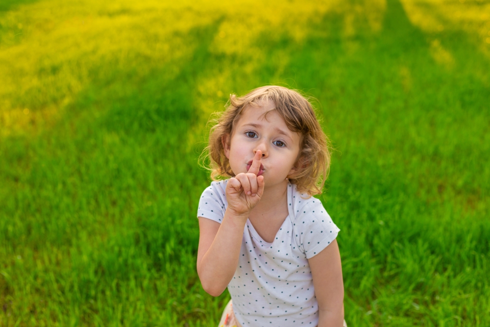 a girl sitting on a green lawn putting her finger over her mouth to indicate “quiet” to the camera.