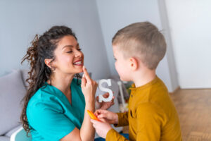 A speech pathologist in green scrubs, holding a wooden letter “S”, pointing to her mouth and interacting with a young boy in a yellow shirt.