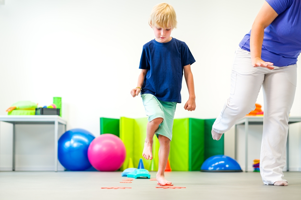 A boy in a blue t-shirt with blond hair lifting his right leg and practicing his balance next to an occupational therapist doing the same thing.
