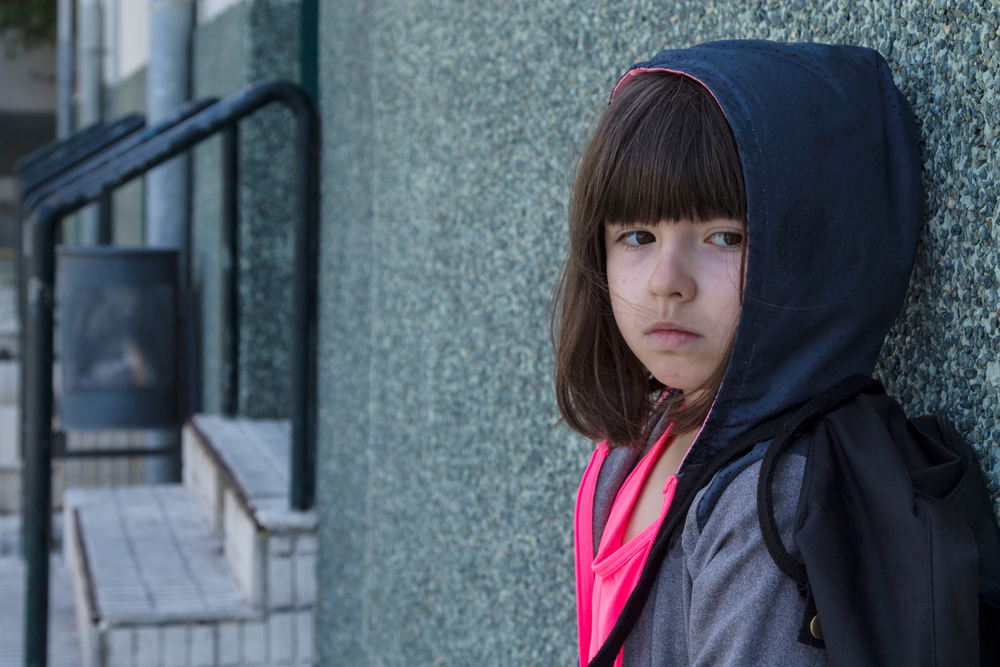 a young girl with chin-length brown hair and bangs, wearing a hooded jacket and a pink shirt underneath, leaning against a wall, looking angry.
