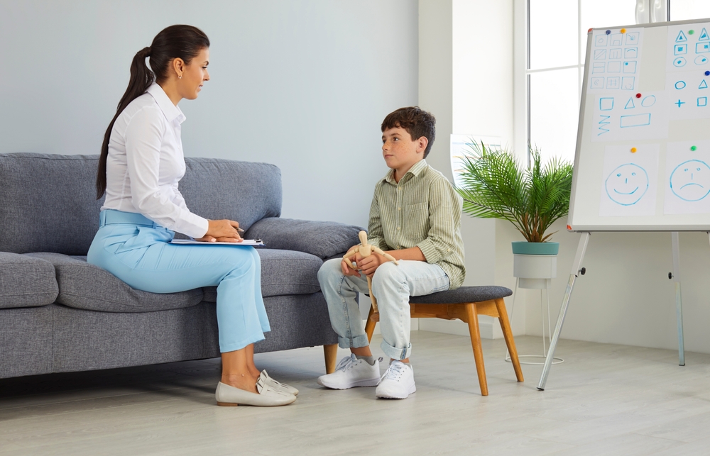 A young boy sitting on a sofa seat, with a dry-erase board behind him and holding a wooden mannequin doll while listening to a therapist speak to him.