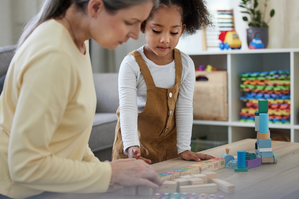 A young black girl wearing overalls standing at a table organizing wooden letter blocks with her therapist in the foreground.