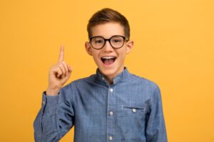 Brown-haired boy with round glasses wearing a denim shirt and enthusiastically indicating he has an idea