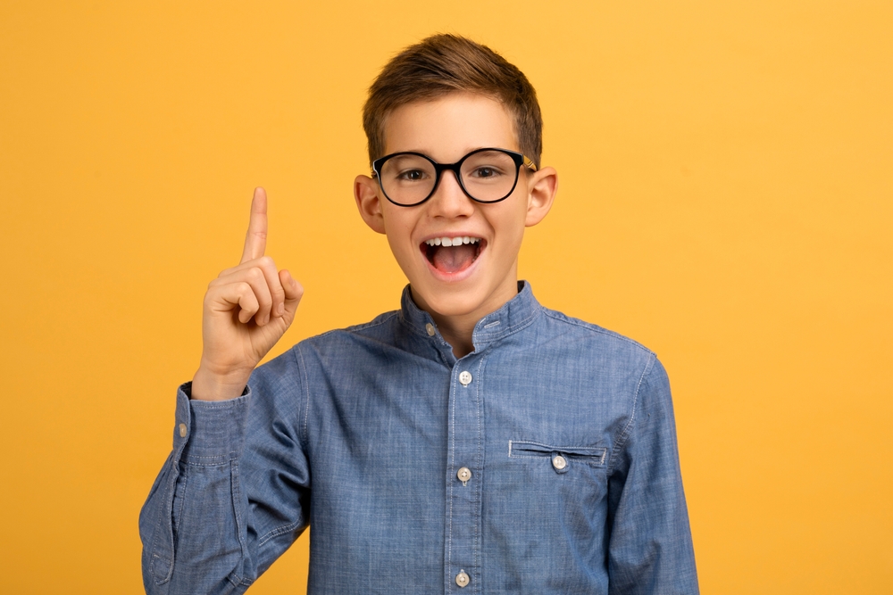 Brown-haired boy with round glasses wearing a denim shirt and enthusiastically indicating he has an idea