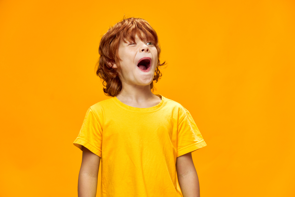 A red-headed child wearing a yellow short-sleeved shirt and looking enthusiastically with his mouth open