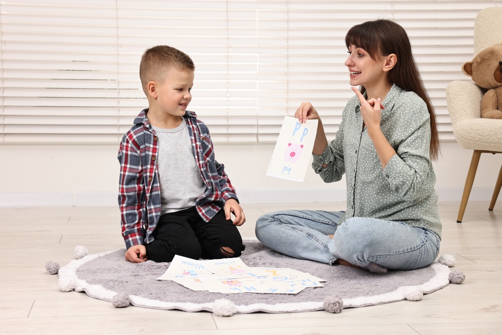 A woman sitting cross-legged on a circular carpet and pointing to her mouth, while a young boy in a plaid shirt watches her.
