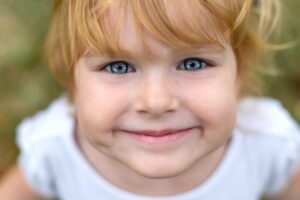 A close-up of a blond-haired child’s smiling face with big blue eyes, wearing a white t-shirt.