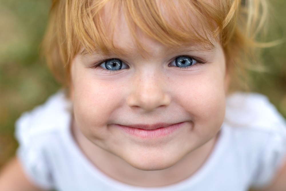 A close-up of a blond-haired child’s smiling face with big blue eyes, wearing a white t-shirt.