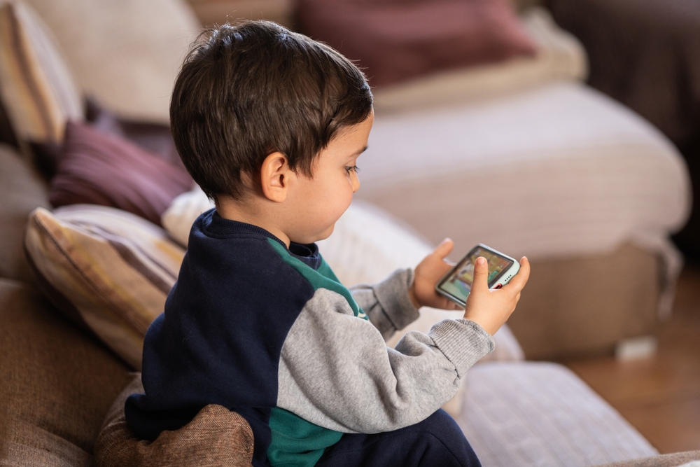 A young child with brown hair sitting on a sofa and using a cell phone to communicate.