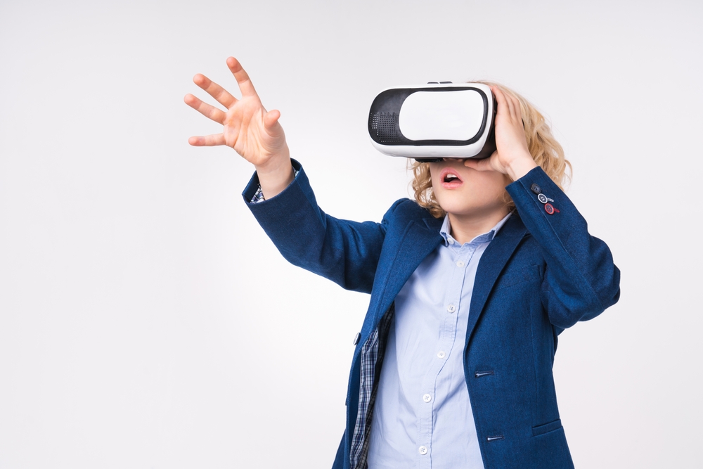  A blond-haired boy wearing a navy blazer and Oxford cloth shirt, reaching his hand out into the air while enjoying a virtual reality experience.