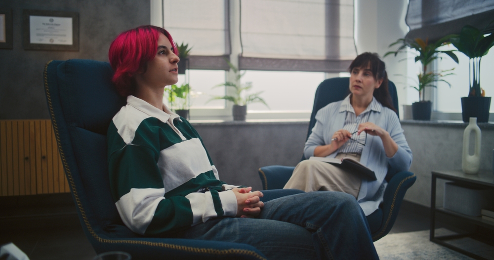 A girl with purple hair wearing a striped rugby shirt and speaking to a therapist who is sitting in the background, listening attentively.
