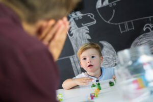 A toddler boy playing with figurines while looking up at an adult man who is making a hand gesture to sleep to him.