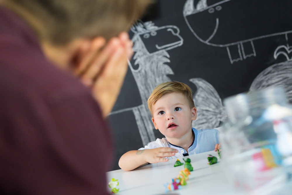 A toddler boy playing with figurines while looking up at an adult man who is making a hand gesture to sleep to him.
