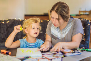 A therapist working with a young blond-haired girl at home, who is holding a yellow highlighter and seated at a table with her.