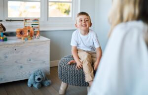 A young boy in a white short-sleeved shirt and khaki pants sitting on a stool, smiling and speaking with a therapist.