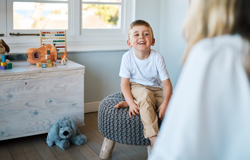 A young boy in a white short-sleeved shirt and khaki pants sitting on a stool, smiling and speaking with a therapist.