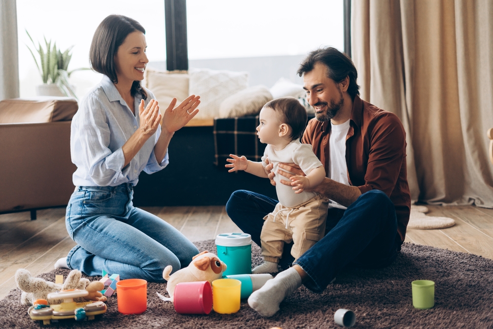 A baby being held up to standing by a man, while a woman is clapping and smiling at the child, surrounded by plastic toys on a large circular rug.