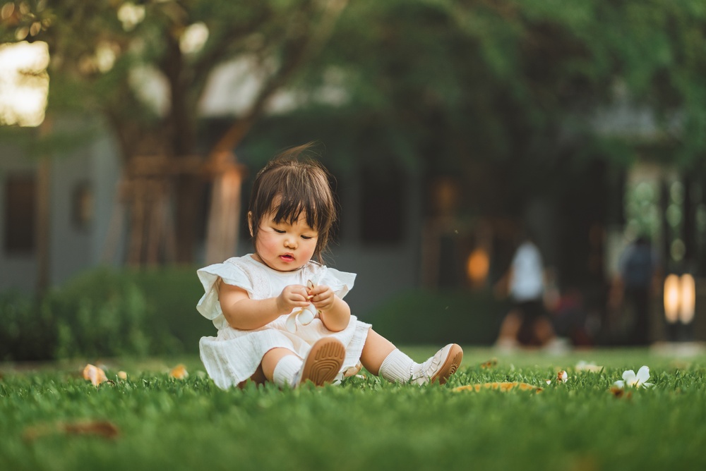 A small asian girl wearing a white dress and shoes sitting on a green lawn and playing with something in her hands.