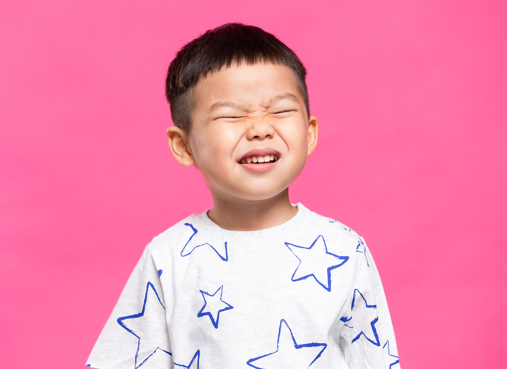 An Asian boy wearing a white shirt with blue stars squinting his eyes closed.