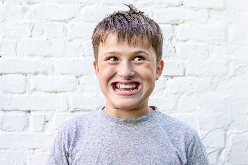 A boy wearing a grey t-shirt, giving an exaggerated smile to the camera.