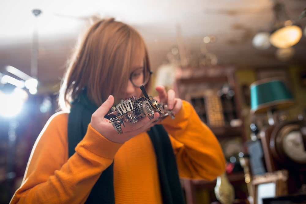A teen wearing an orange shirt and scarf with long hair and glasses staring intently at a model car.