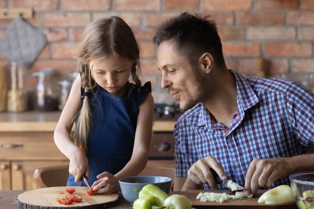 A father looking on as his young daughter cuts baby tomatoes on a cutting board while standing in the kitchen.