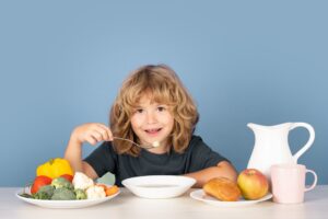 Brown-haired child sitting at a table and enjoying a spoonful of soup with plates of fruits and vegetables surrounding her.