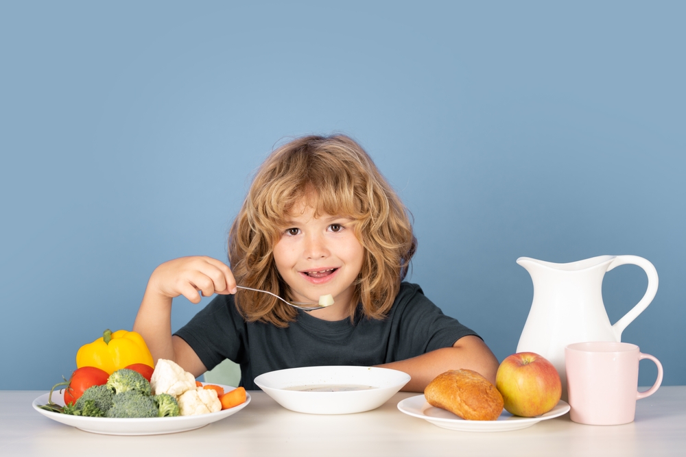 Brown-haired child sitting at a table and enjoying a spoonful of soup with plates of fruits and vegetables surrounding her.