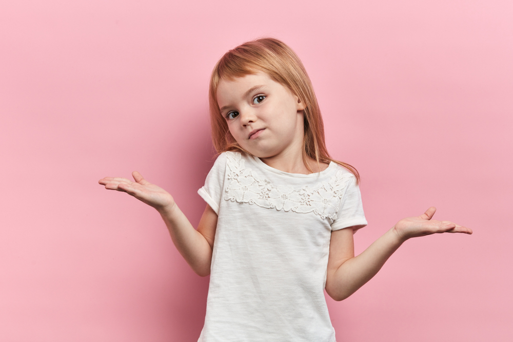 A young blond-haired girl wearing a white t-shirt, shrugging her shoulders and turning her palms up.
