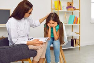 A female therapist with a clipboard gently placing her hand on the head of a young girl who is sitting with her face in her hands during a therapy session in a calm, child-friendly office.