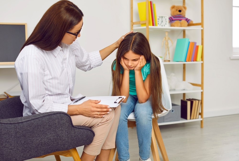 A female therapist with a clipboard gently placing her hand on the head of a young girl who is sitting with her face in her hands during a therapy session in a calm, child-friendly office.