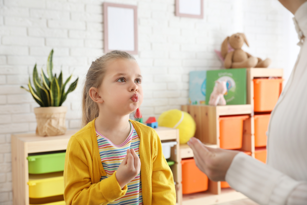 A blond-haired girl with a long ponytail speaking with an adult while puckering her lips.
