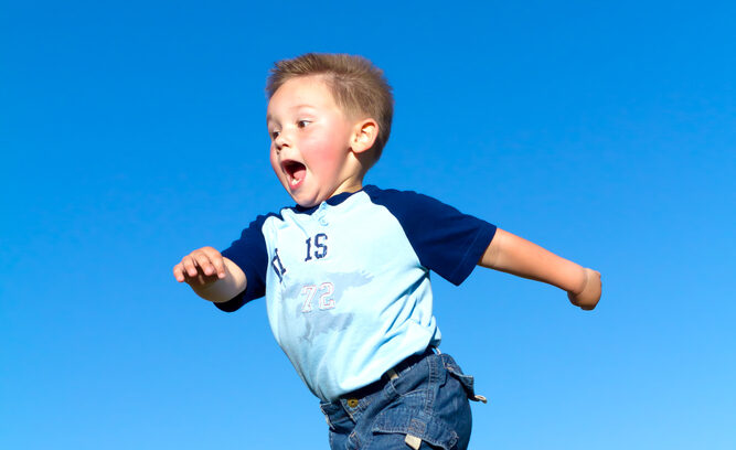 A young boy wearing shorts and a t-shirt running in a field with flushed cheeks and mouth wide open.