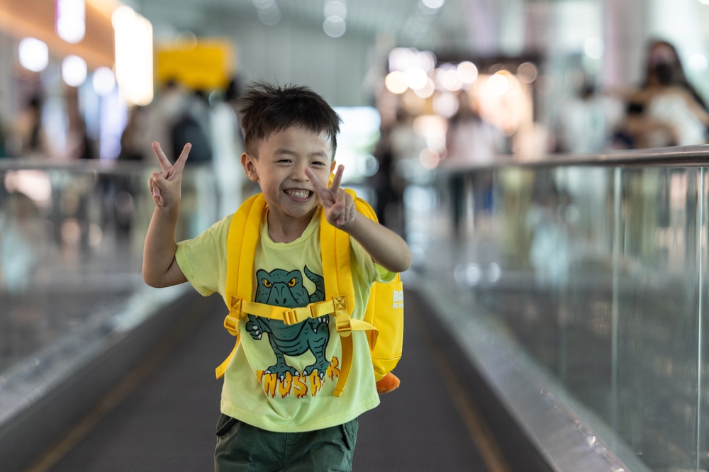 An Asian child making a peace sign with both sets of fingers as he’s running down a moving walkway, wearing a yellow backpack and dinosaur shirt.
