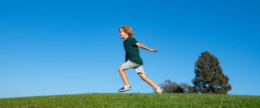  A young boy in a green shirt and khaki shorts running in a field with his arms open and a treat in the background.