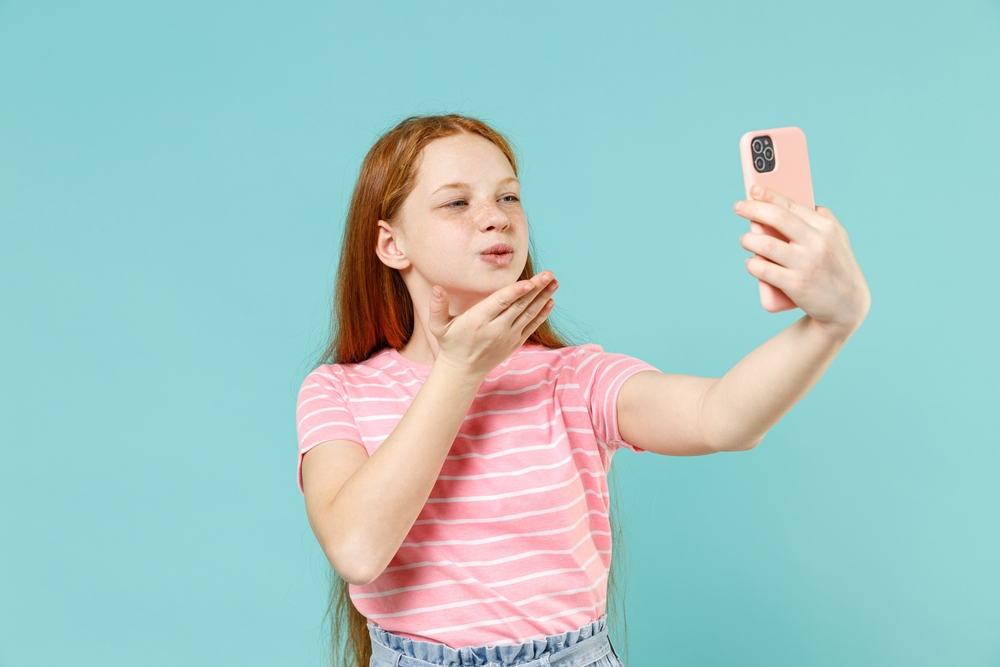 A tween with long red hair and a pink striped shirt taking a selfie of herself and blowing a kiss.