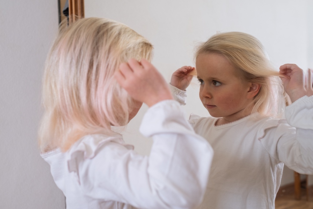 young blond-haired girl wearing a white top, touching her hair and admiring herself in the mirror. 