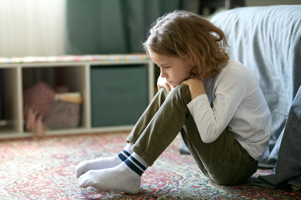 A young boy with shoulder-length hair sitting by his bed on the carpet, resting his chin on his knees.