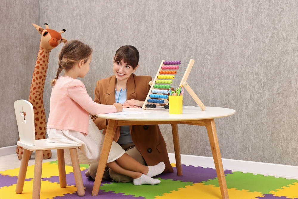 A therapist wearing a brown blazer, smiling and kneeling by the table where a young girl in a pink sweater and dress is working.