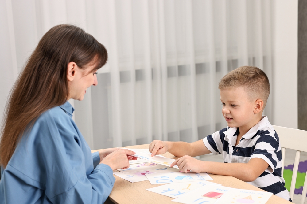 Long brown-haired therapist working at a table with a boy wearing a blue and white striped T-shirt.