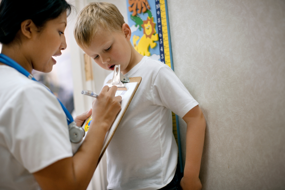 Healthcare professional measures a young child's height during a pediatric evaluation while recording information on a clipboard in a medical exam room.