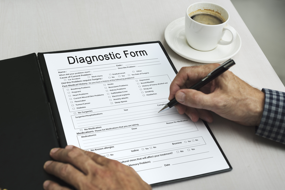 Person completes a medical diagnostic form on a clipboard while seated at a desk with a cup of coffee nearby. 