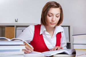 A young woman studies at a desk surrounded by stacks of textbooks, taking notes with a pen.