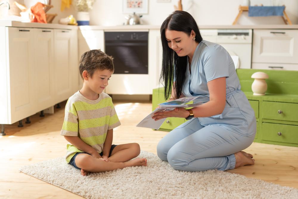 ABA therapist in scrubs kneeling on living room rug, reading a book with young boy during in-home ABA therapy session